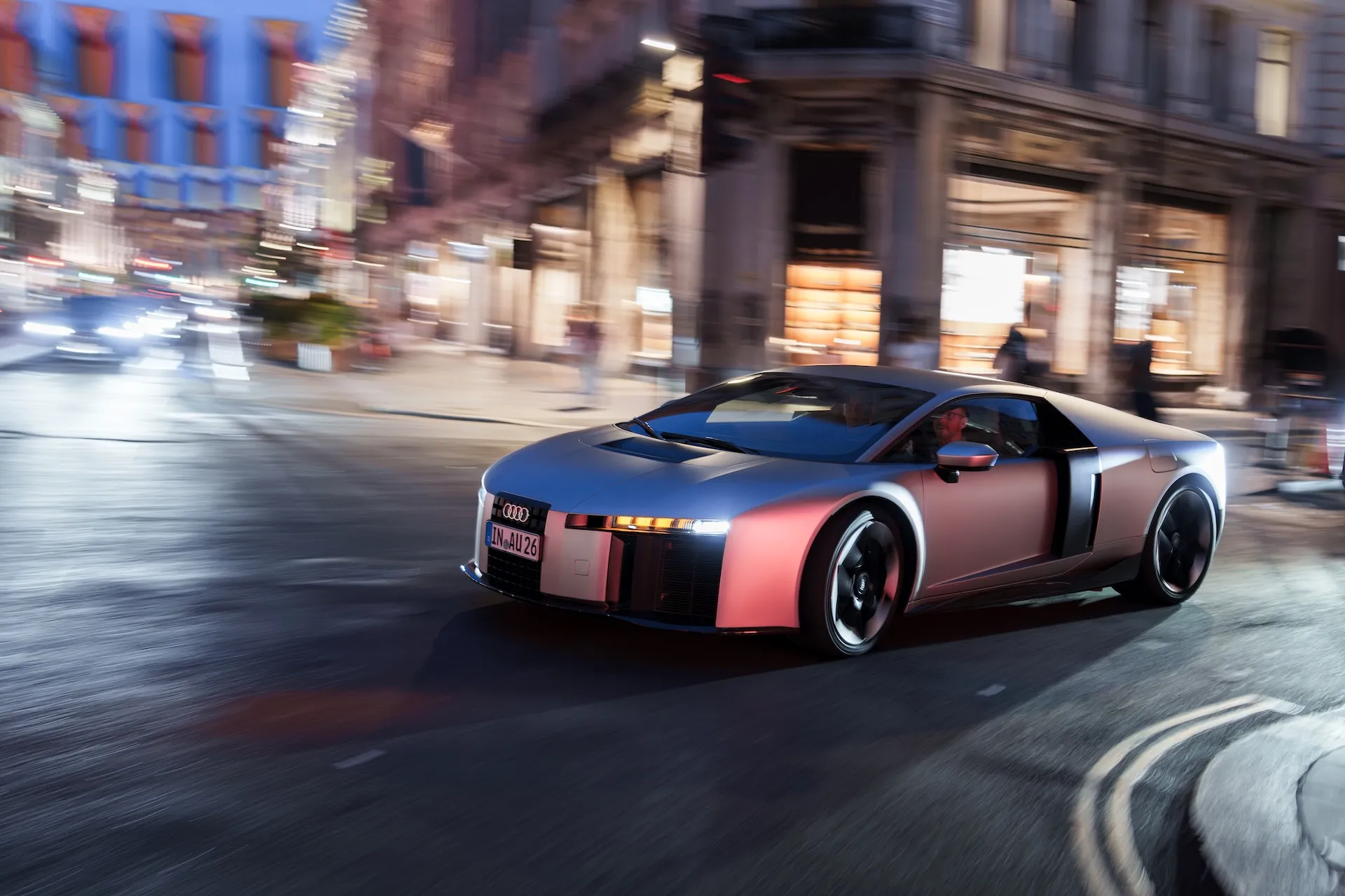Audi Nuvolari hybrid supercar in Titanium and pink dual-tone finish driving through central London at dusk, with motion-blurred storefronts in the background.