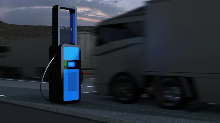 -charging i-light heavy-duty EV charger with illuminated blue panels and transparent display, positioned roadside as trucks pass at dusk