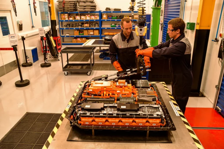 Two technicians in safety glasses and insulated gloves dismantle a high-voltage EV battery pack on a workbench at an Autocraft REVIVE EV battery remanufacturing workshop