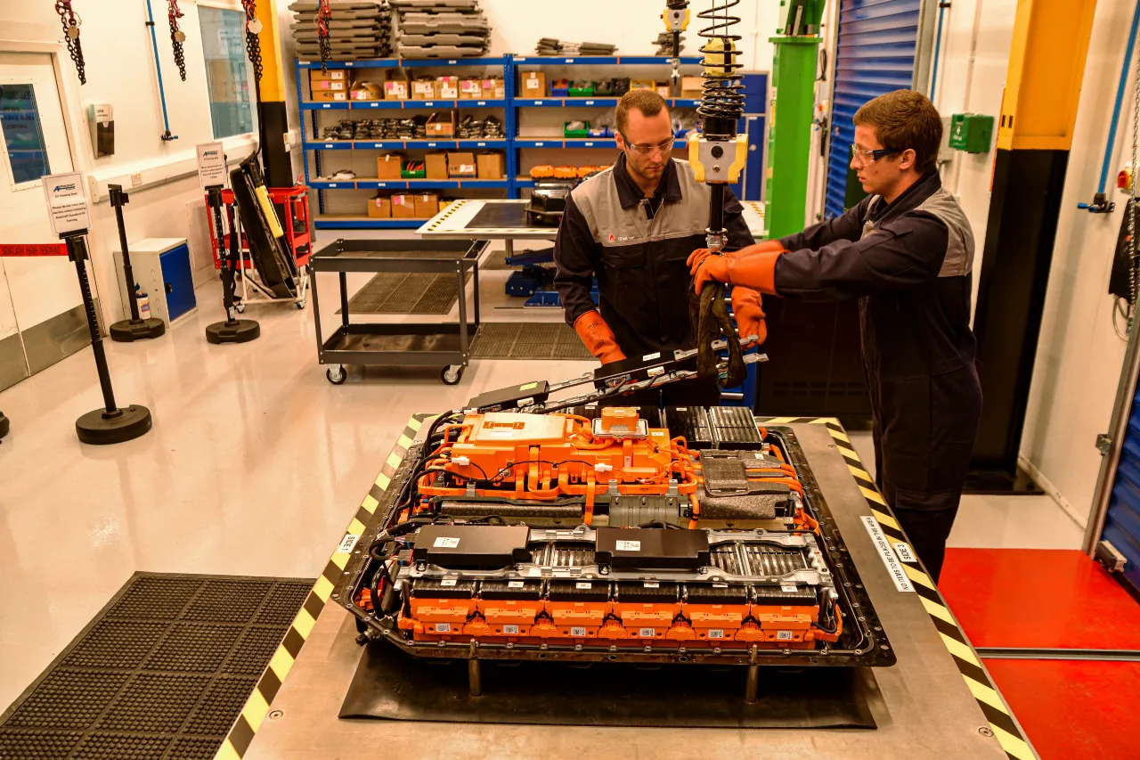 Two technicians in safety glasses and insulated gloves dismantle a high-voltage EV battery pack on a workbench at an Autocraft REVIVE EV battery remanufacturing workshop